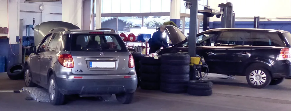 Zwei Fahrzeuge mit geöffneter Motorhaube in der Werkstatthalle von Auto Wittmann, dazwischen ein Mitarbeiter am Motorraum und gestapelte Reifen